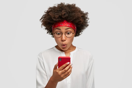attractive-curly-dark-skinned-female-with-afro-hairstyle-looks-agitated-at-screen-of-smart-phone-impressed-by-message-content-recieved-from-friend-has-surprised-facial-expression-stands-indoor
